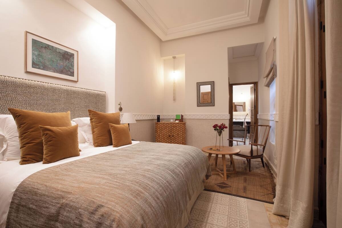 A hotel room with a large bed, a small round table, a chair, and a view of the bathroom and kitchen through an open door.
