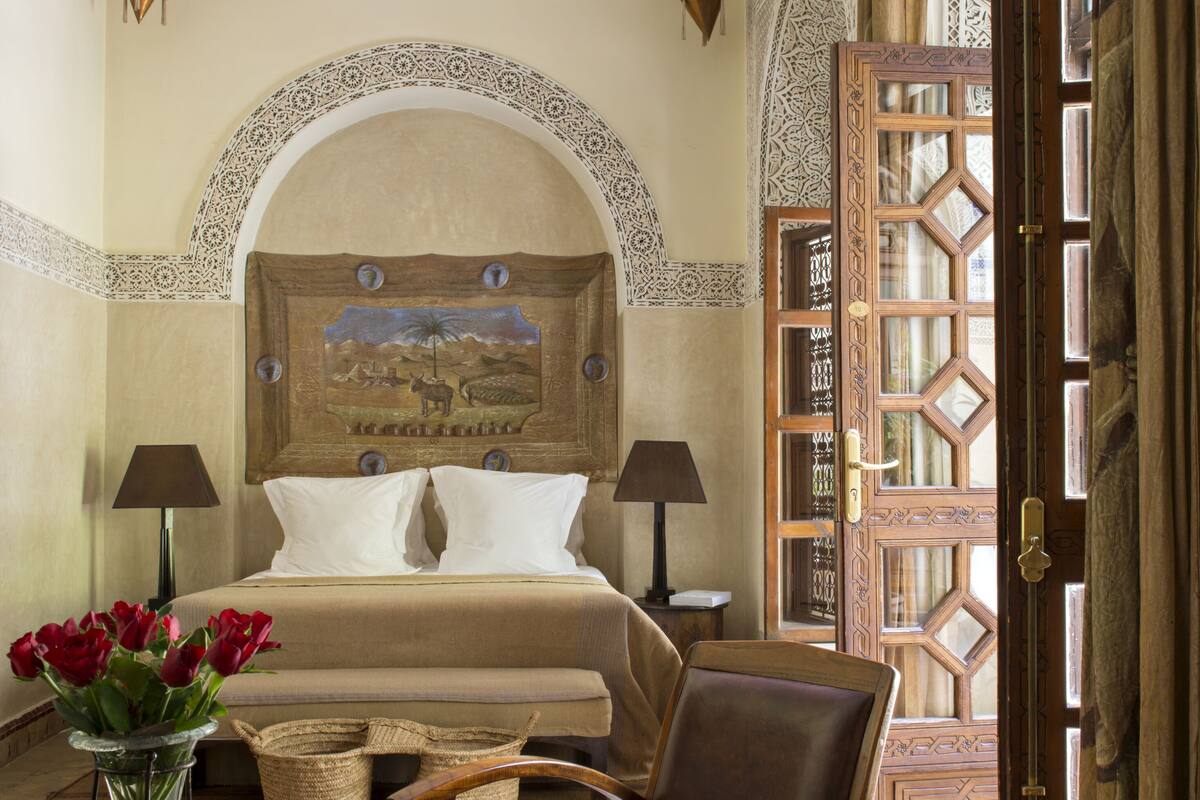 A bedroom with a large bed, two bedside tables, a chair, a vase of red flowers, and a decorative door.