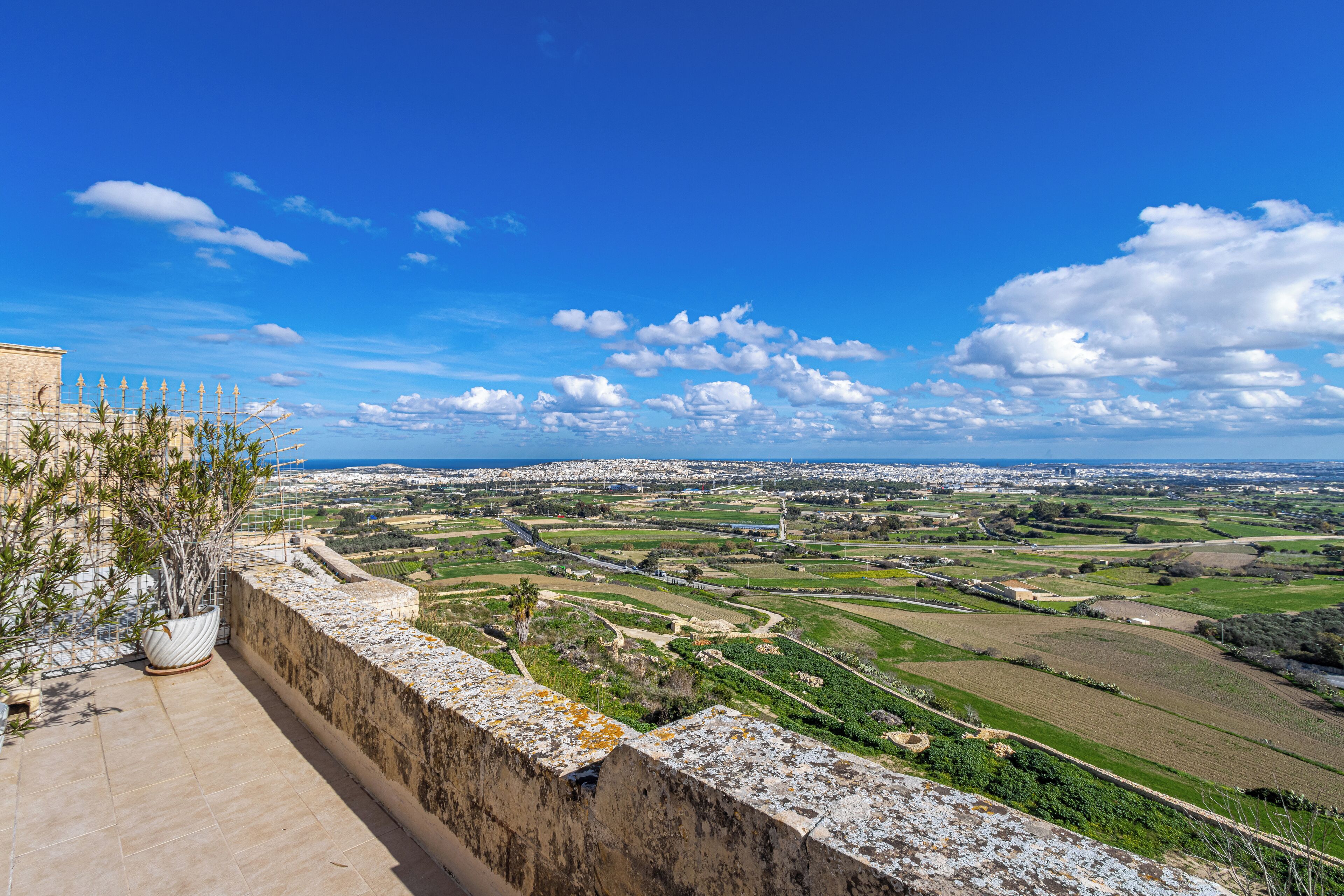 A balcony with a view of a coastal landscape, including fields and a distant cityscape.