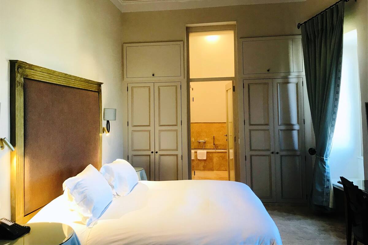 Quarto de hotel com uma cama, uma escrivaninha com telefone, um espelho, uma porta que dá para um banheiro e uma cortina.