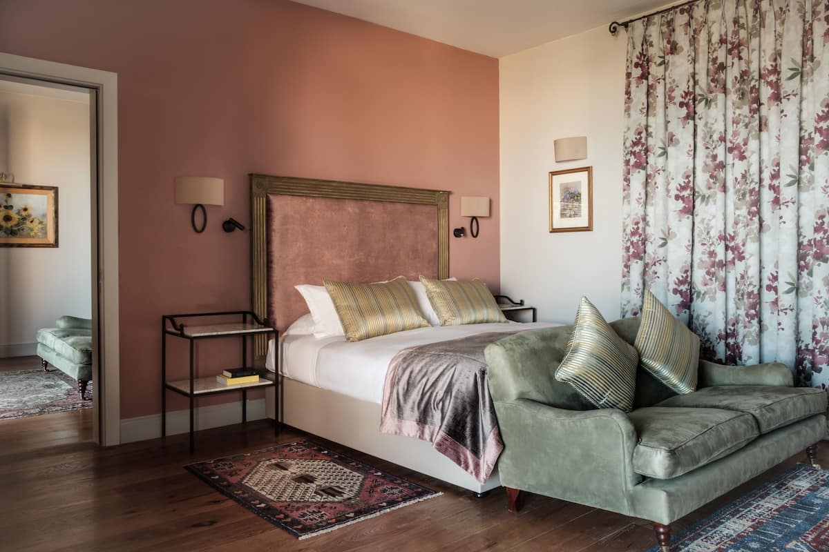 Quarto com cama, poltrona verde, mesa de cabeceira, abajur e cortina florida.