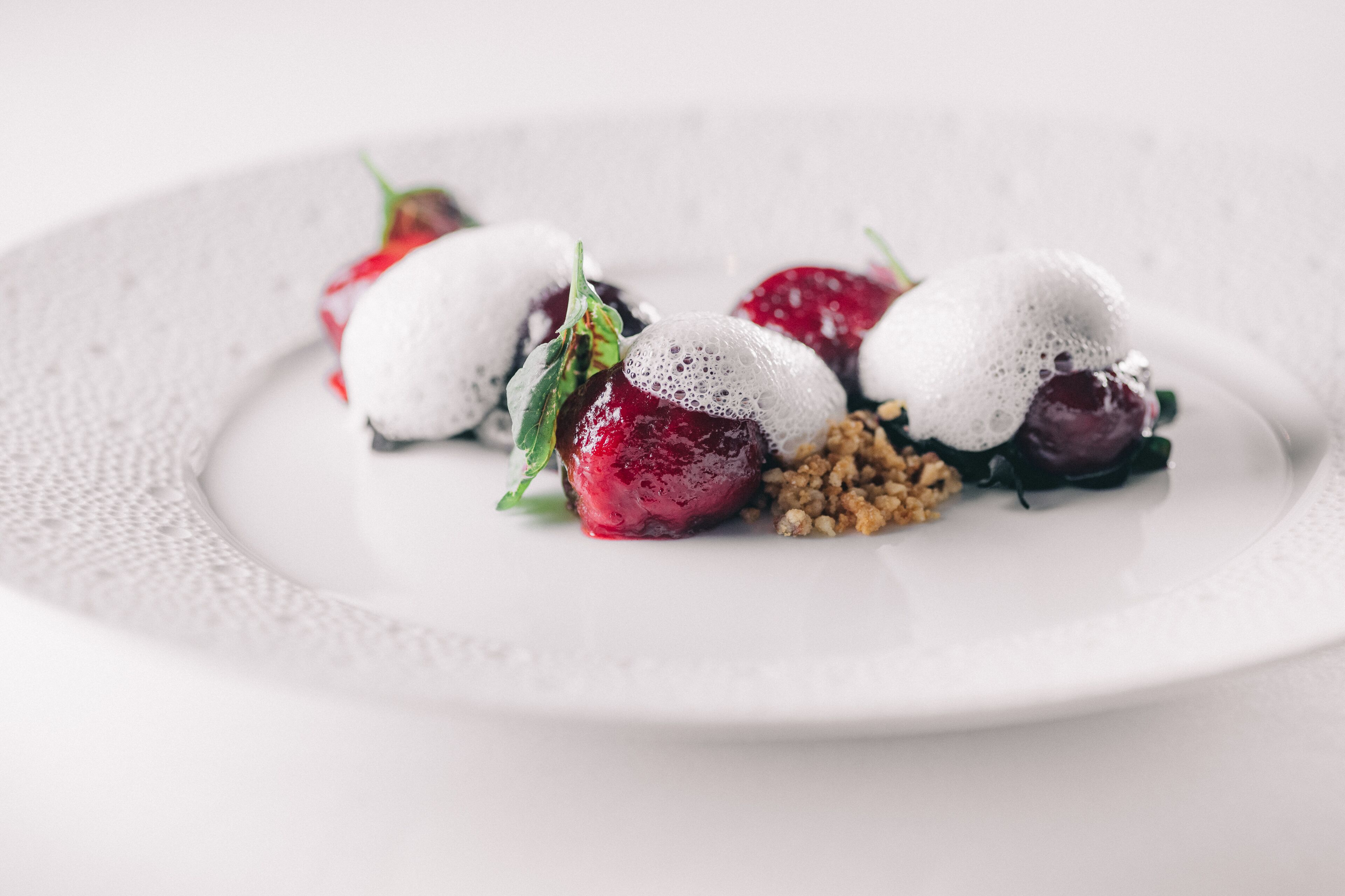 Um prato branco com três morangos cobertos por espuma branca e decorado com folhas verdes e granola.