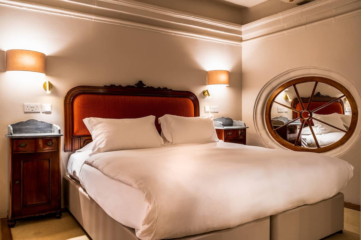 Uma cama bem arrumada com lençóis brancos, duas luminárias de cabeceira, uma mesa de cabeceira de madeira e uma janela redonda com moldura de madeira.