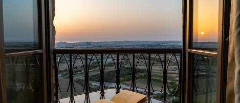 A balcony with a table holding an open book, overlooking a landscape with a sunset.