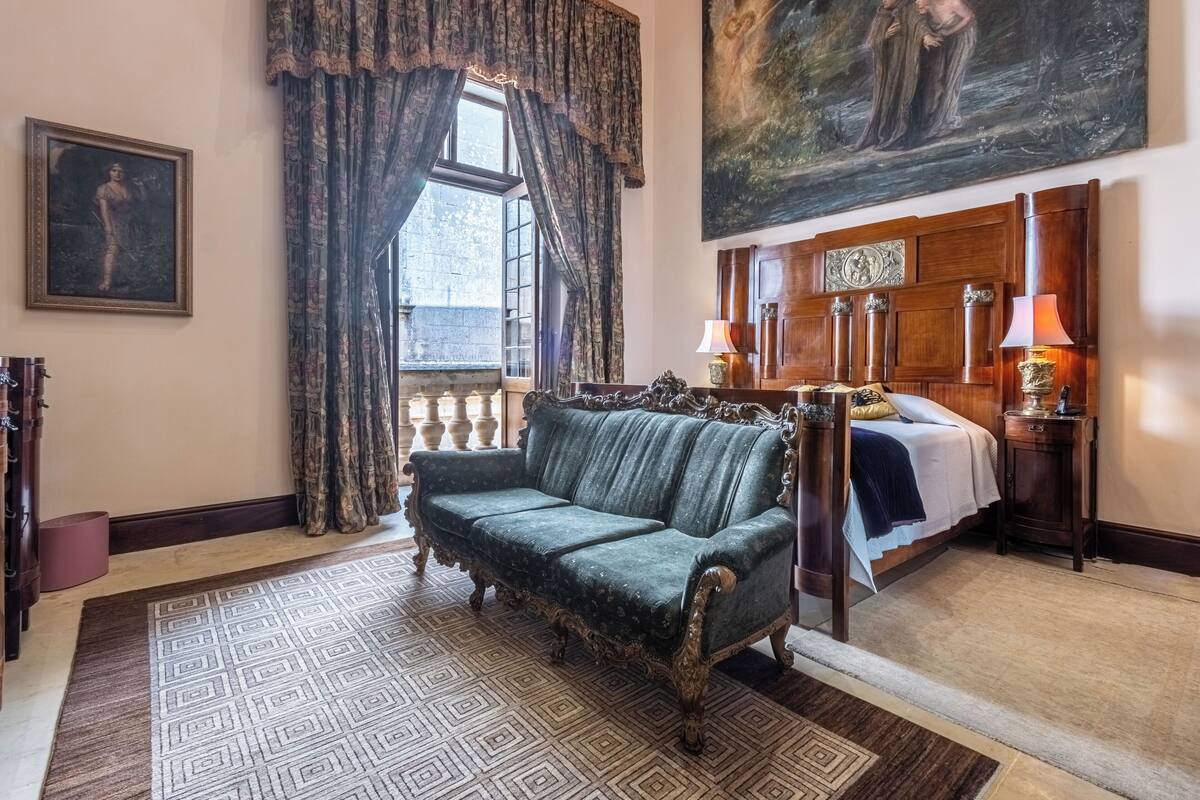 Quarto com um sofá verde antigo, uma cama com colcha branca e azul, um quadro grande na parede e um tapete estampado.