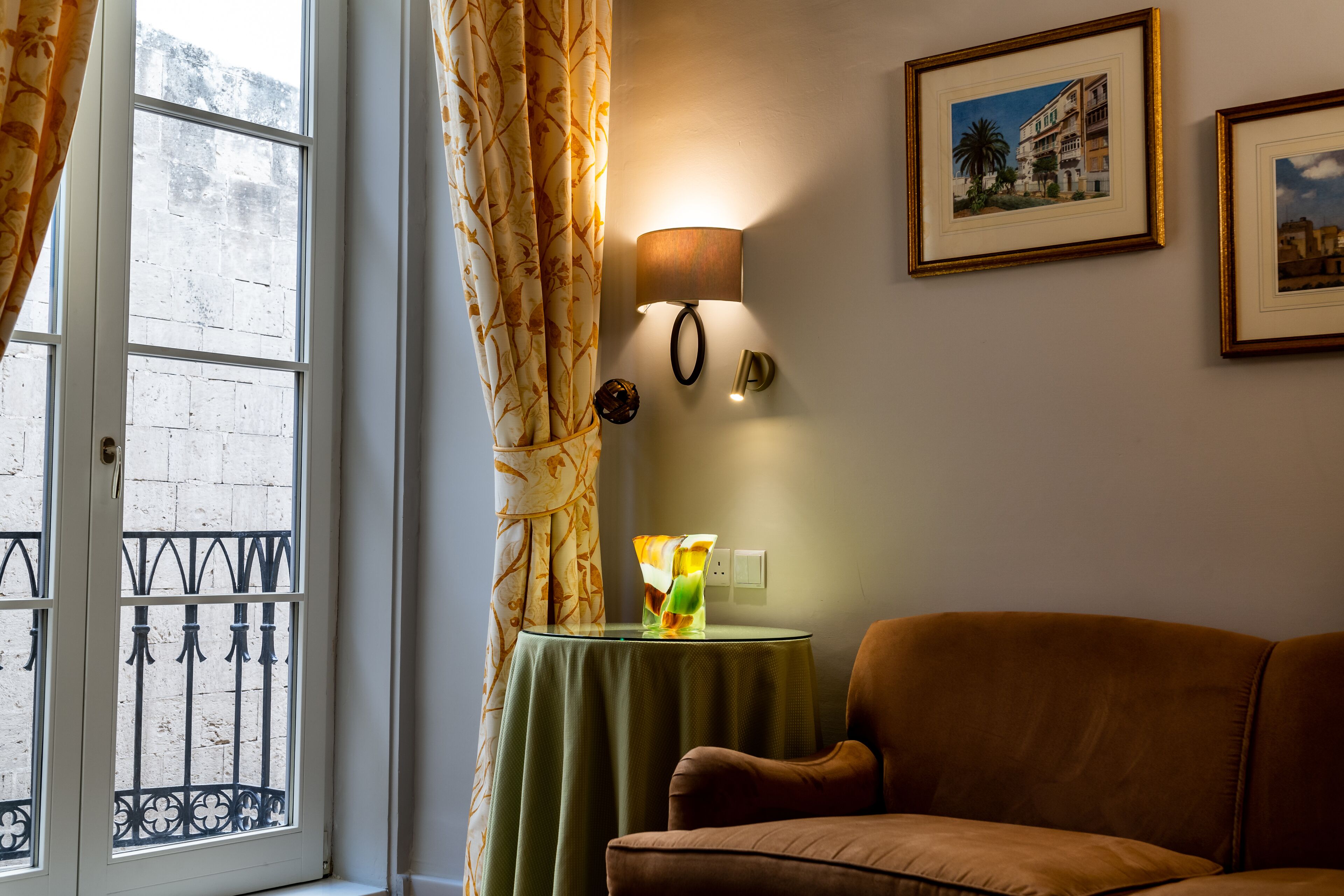 Una habitación con ventana, una mesa verde con lámpara, un sofá marrón y dos cuadros en la pared.