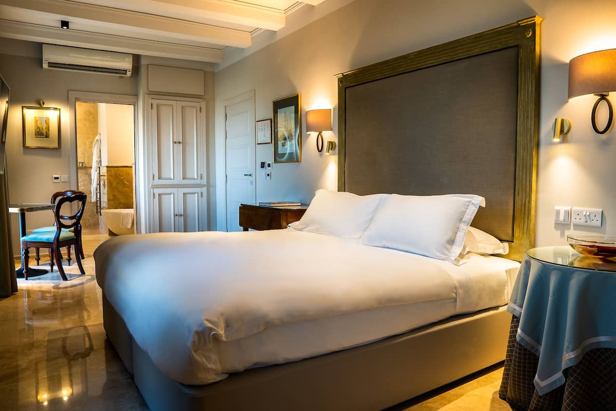 Quarto de hotel com uma cama grande, uma cadeira, uma mesa e um banheiro visível por uma porta aberta.