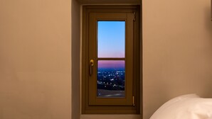 A room with a bed, a window with a view of a cityscape at dusk, and a door with a handle.