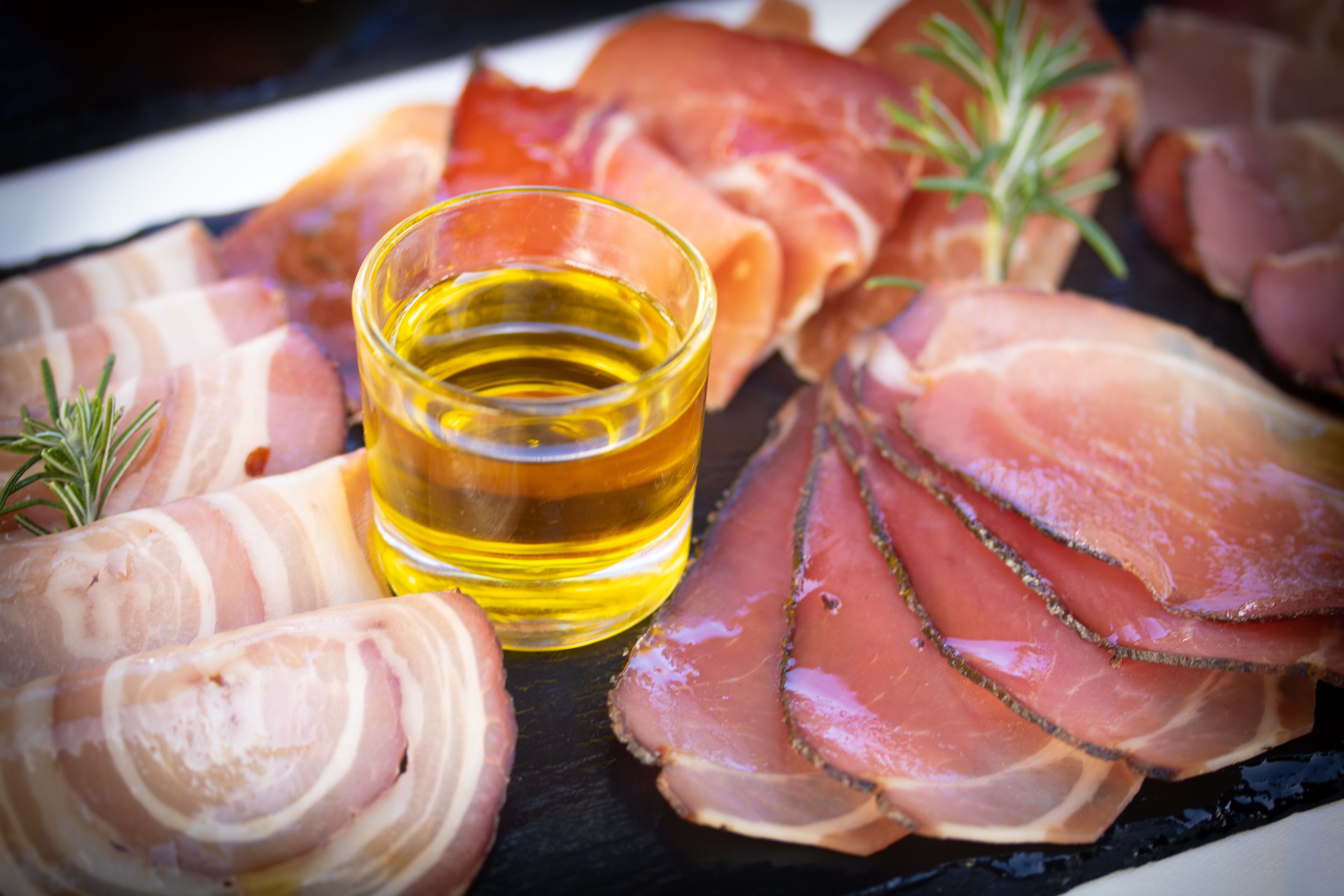 A platter of prosciutto and bacon slices with a glass of oil and rosemary sprigs.