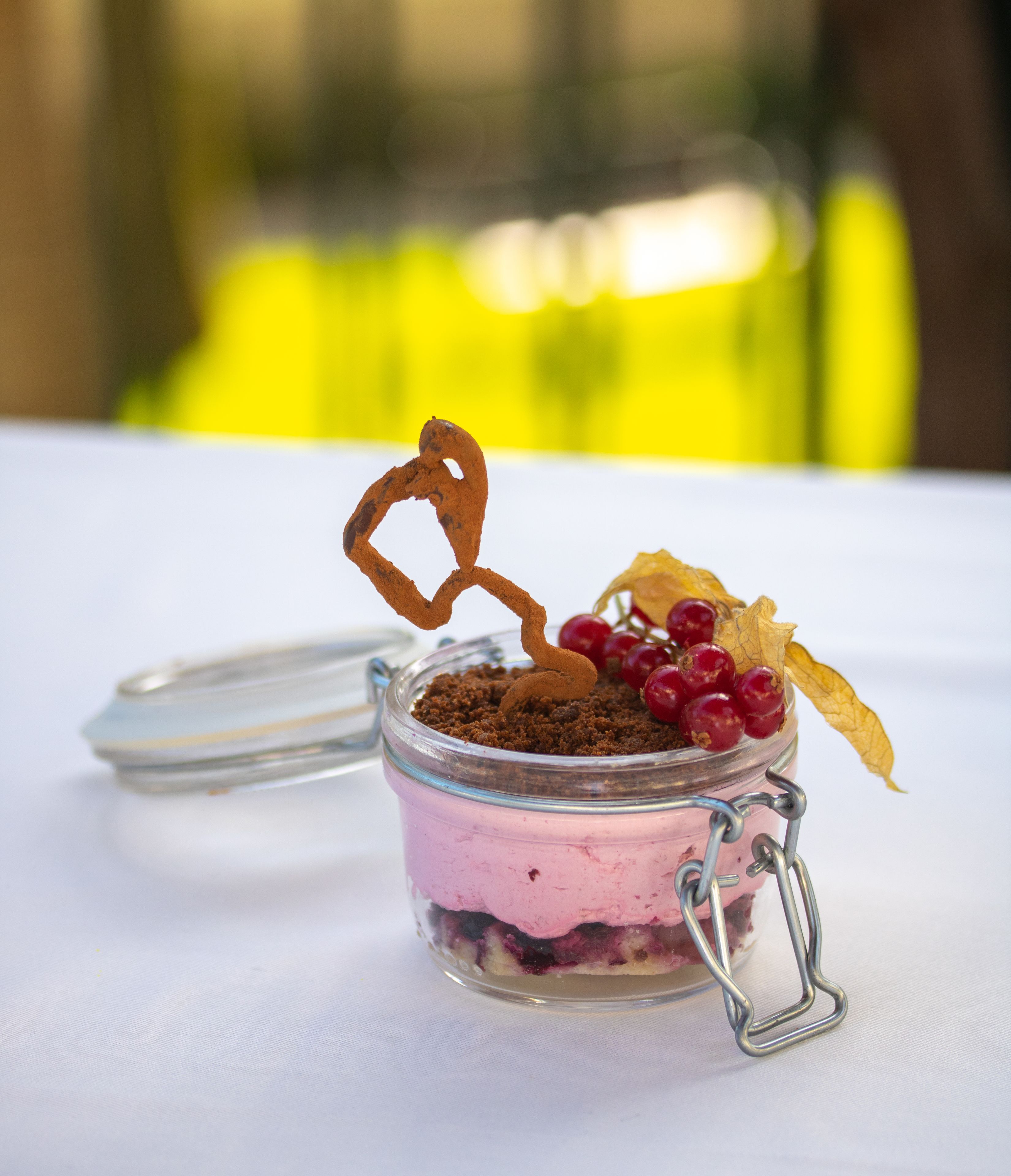 Un dessert servi dans un bocal en verre muni d’un fermoir métallique, surmonté d’une décoration en chocolat en forme de cœur, de quelques baies rouges et d’une feuille séchée.