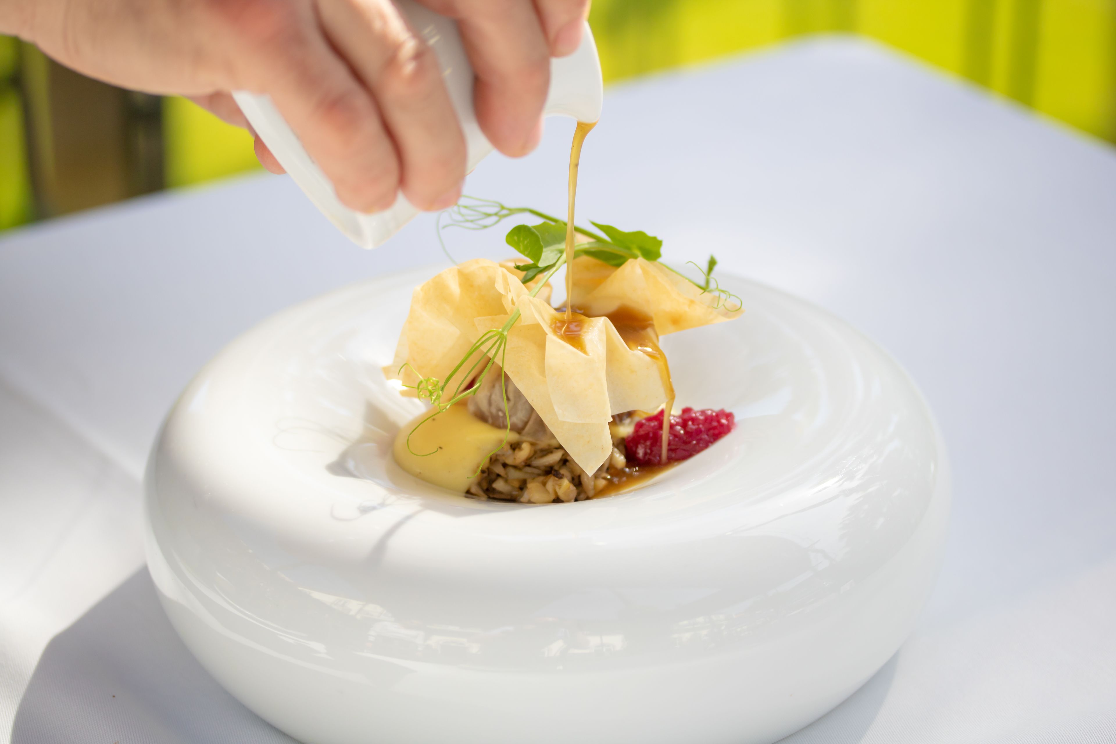A white ceramic bowl with a dessert that includes a pastry, nuts, and a sauce being poured over it.