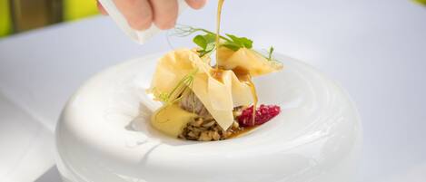 A white ceramic bowl with a dessert that includes a pastry, nuts, and a sauce being poured over it.