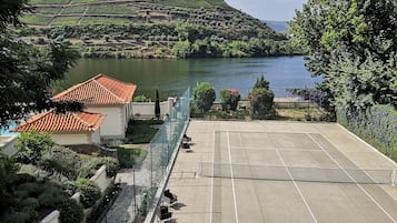 A tennis court with a net, surrounded by a fence and benches. In the background, there is a vineyard and a body of water.