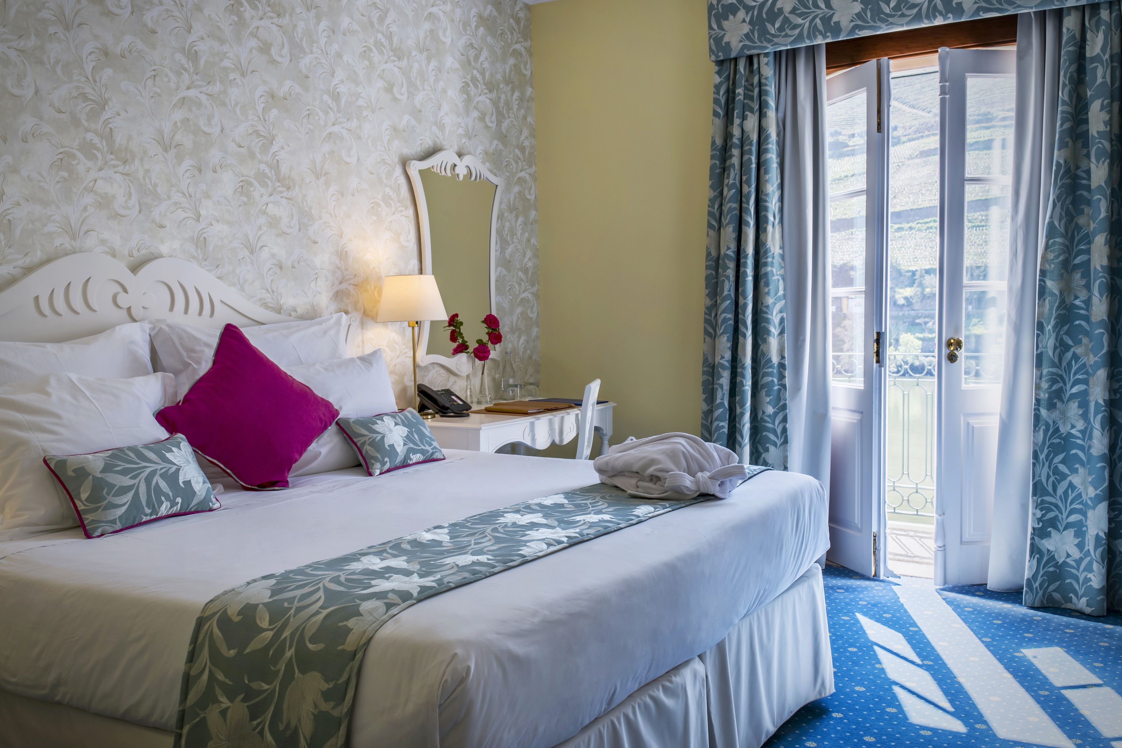 A hotel room with a bed, a desk, a mirror, and a view of the outdoors.