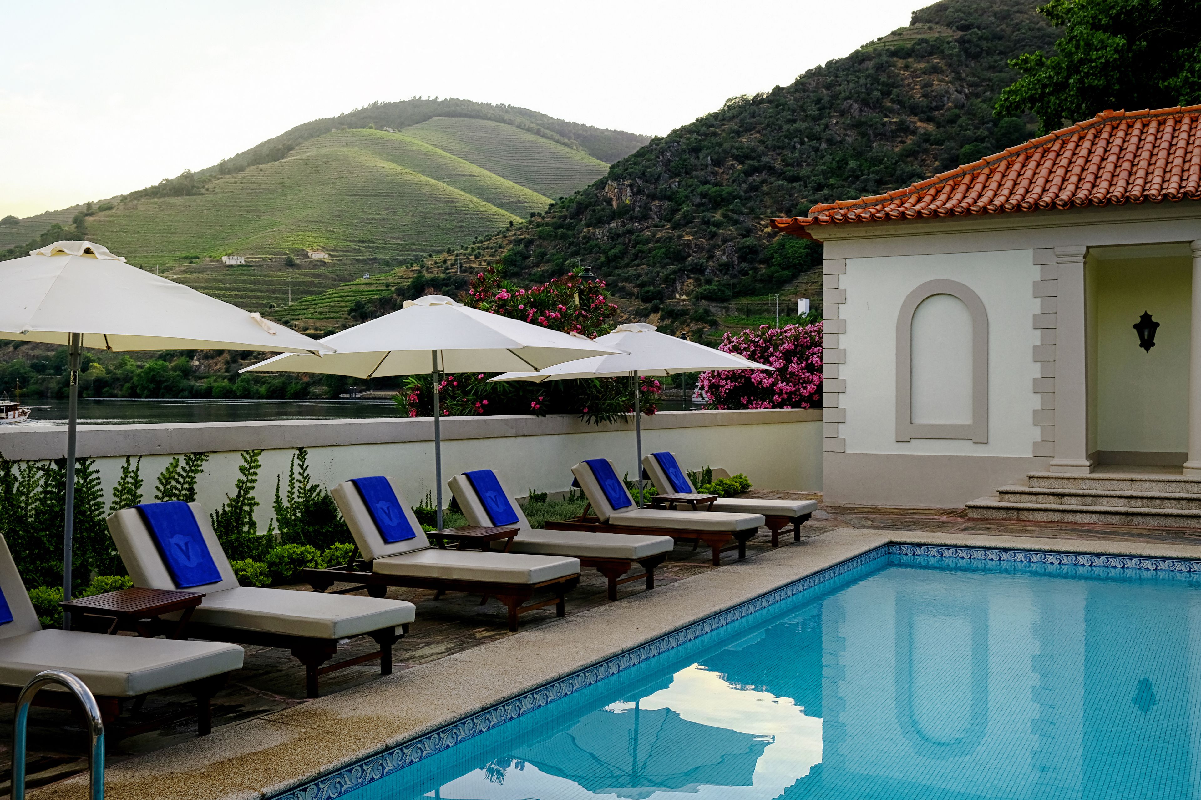 A pool area with lounge chairs and umbrellas, a small building, and a view of green hills.