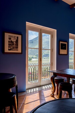 A room with a large window overlooking a scenic landscape, a table with chairs, and framed pictures on the wall.