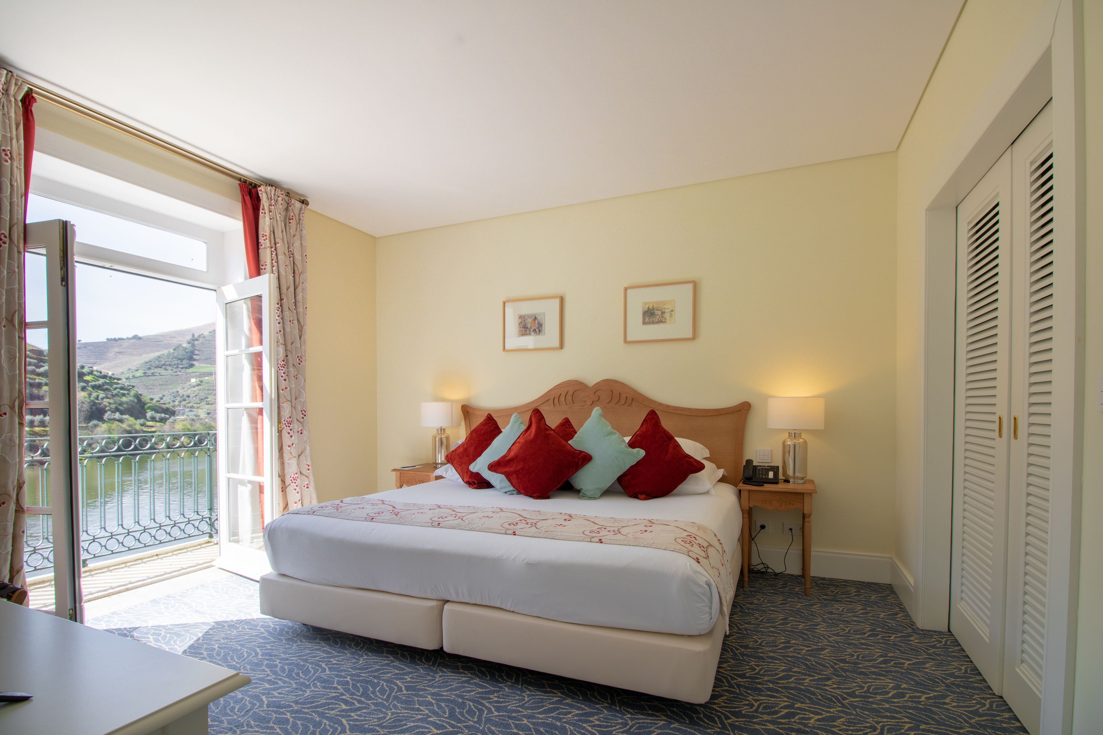 A hotel room with a bed, two bedside lamps, a nightstand, a window with curtains, and a door with shutters.