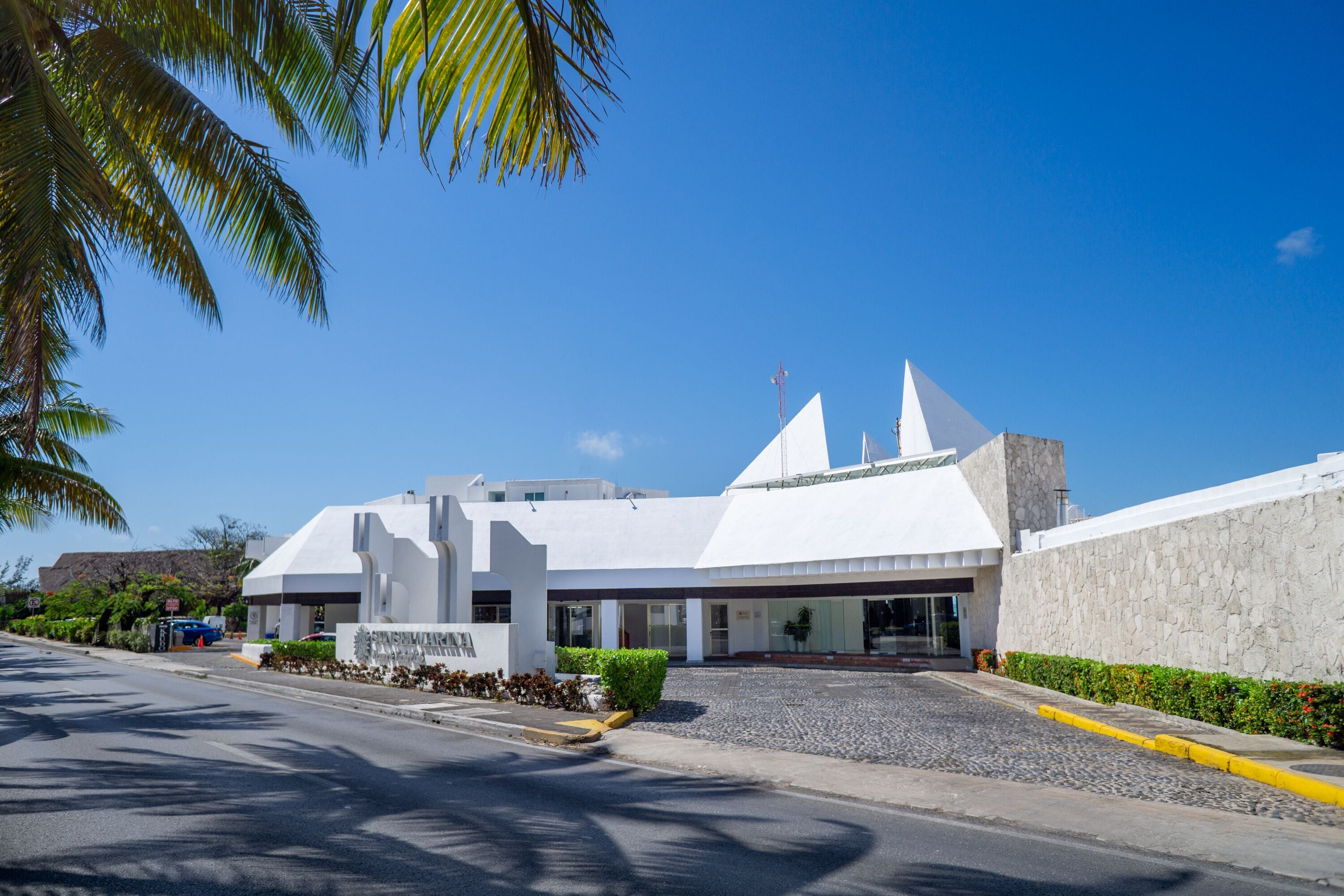 A modern building with a white facade and a sloped roof, featuring a prominent entrance and a landscaped area in front.