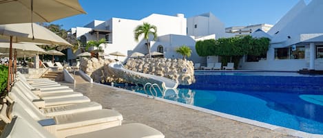 Un espace aménagé au bord de la piscine, avec des chaises longues et des parasols, une piscine à l’eau d’un bleu limpide et des bâtiments blancs au design moderne.