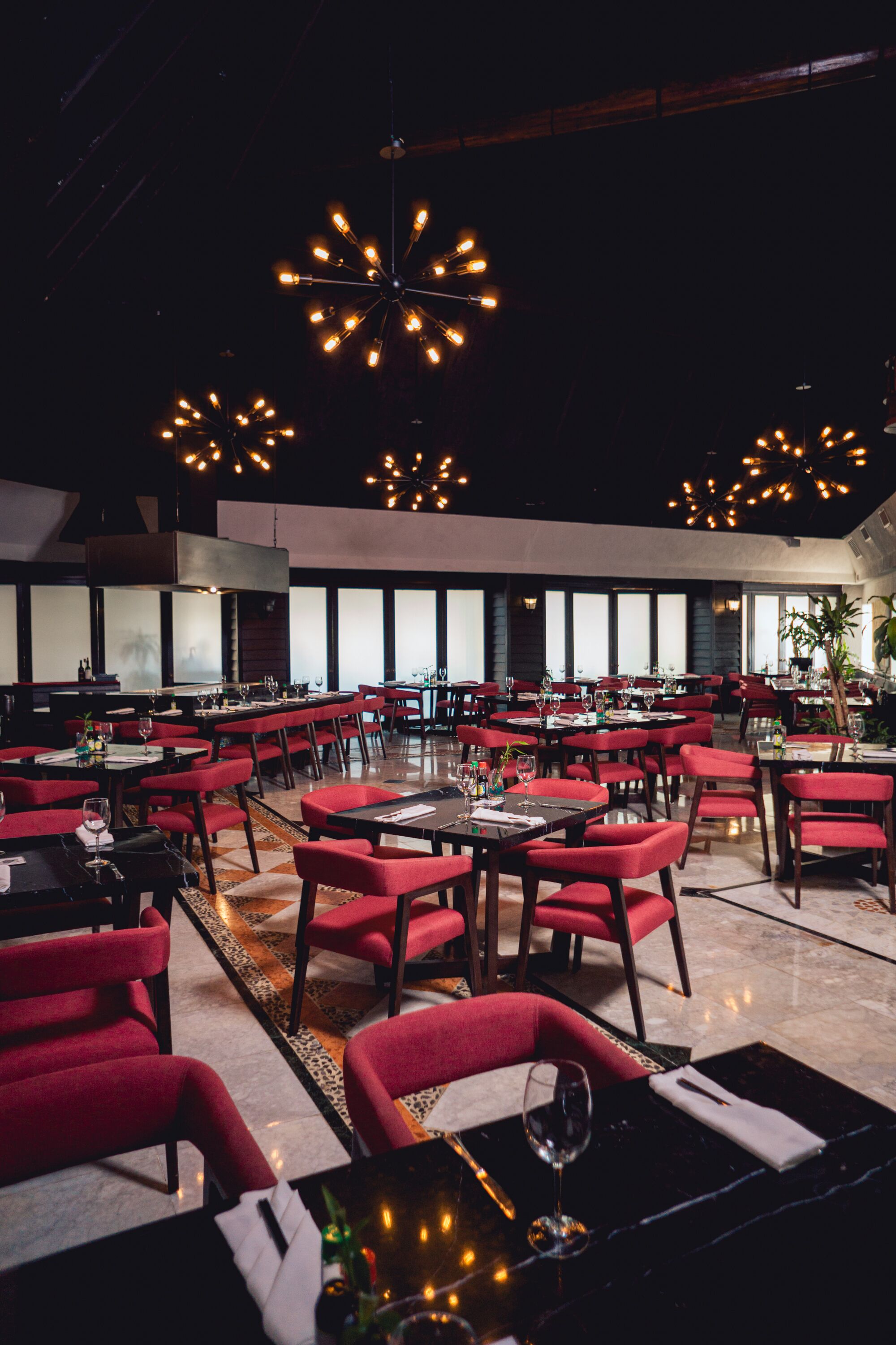 A restaurant with multiple tables and red chairs, featuring a modern chandelier and a large window in the background.
