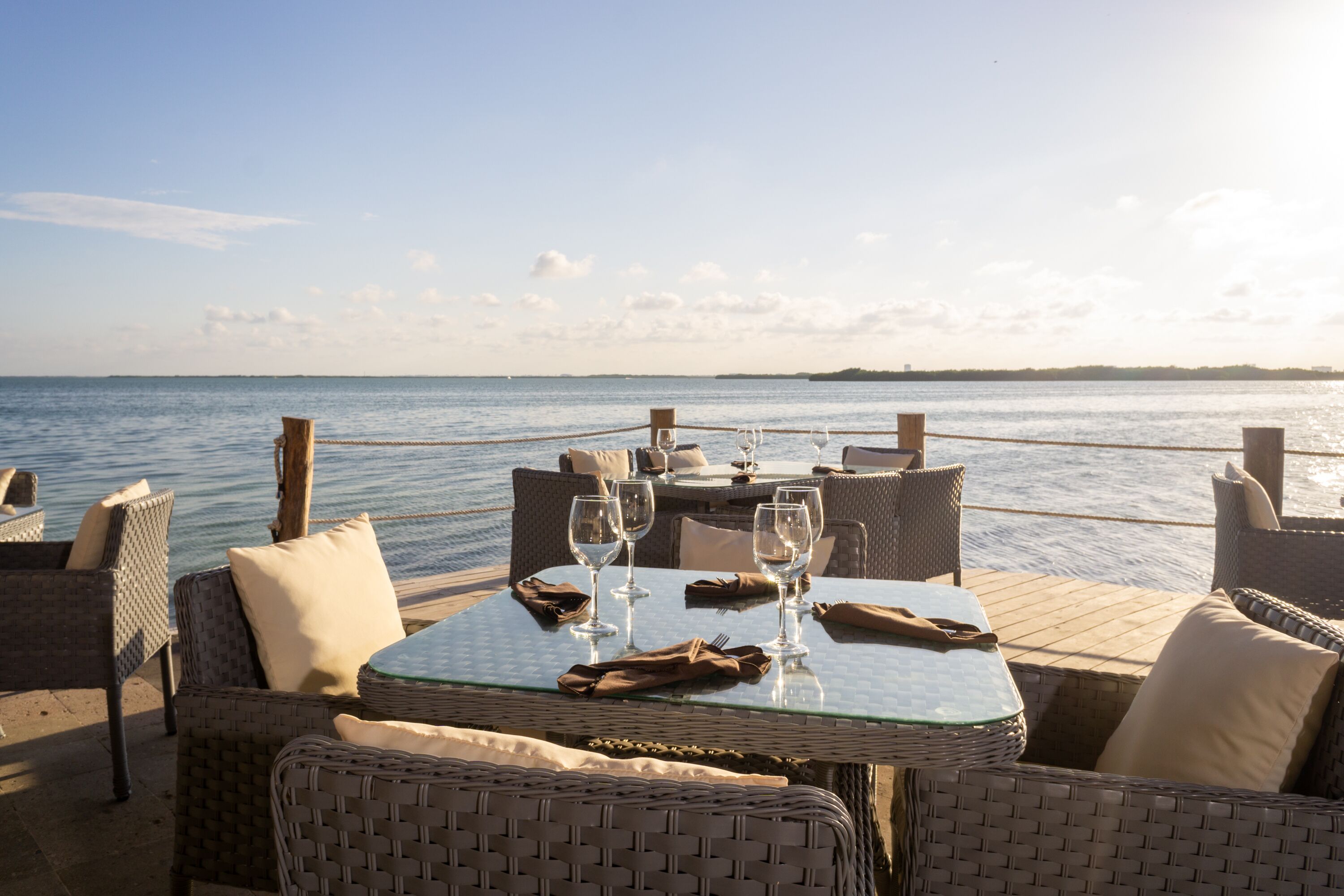 A seaside dining area with wicker furniture, glass tables, and wine glasses, overlooking the sea.