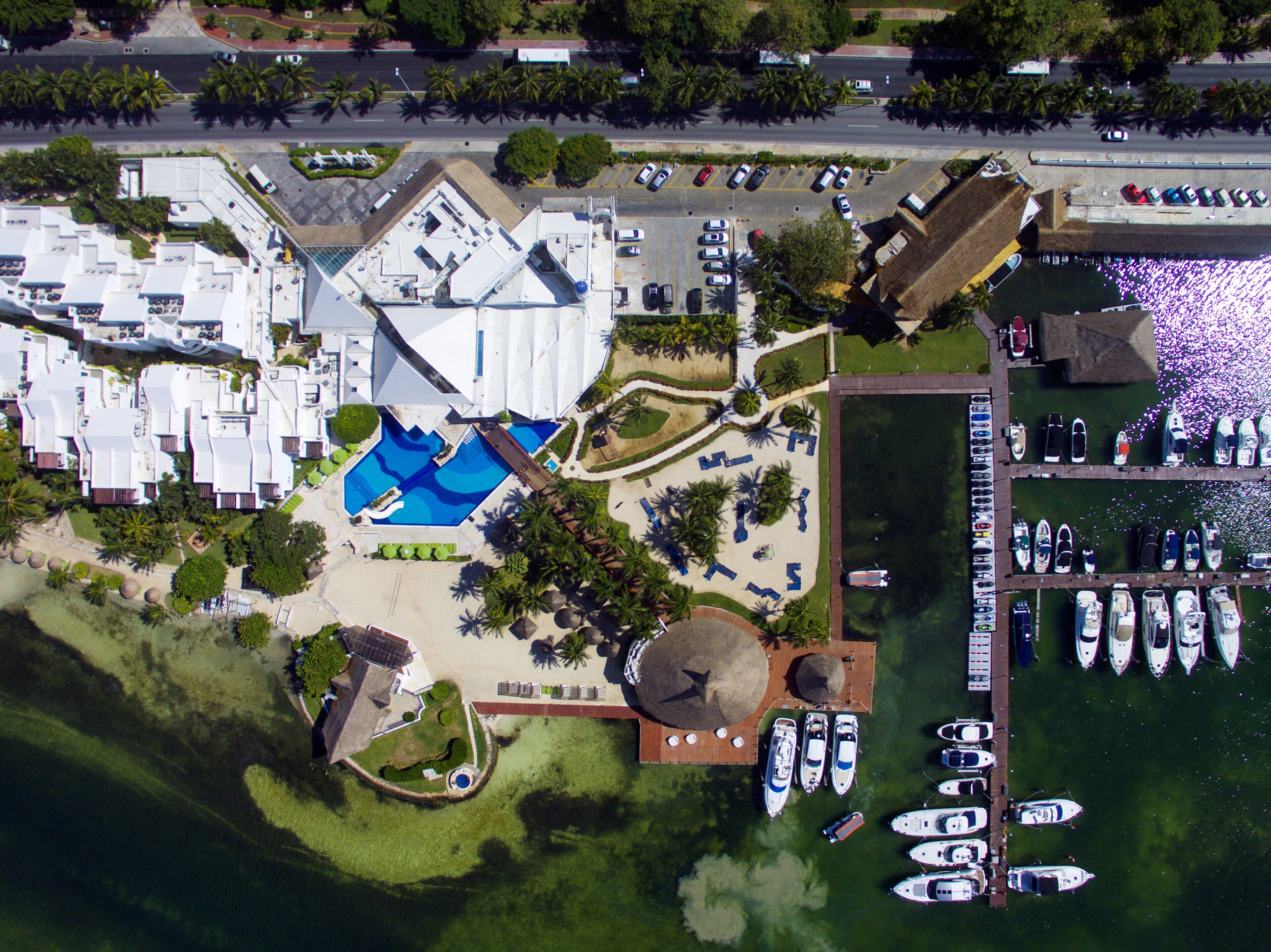 Vista aèria d'un complex turístic amb una piscina, un port esportiu amb vaixells i una zona d'aparcament.
