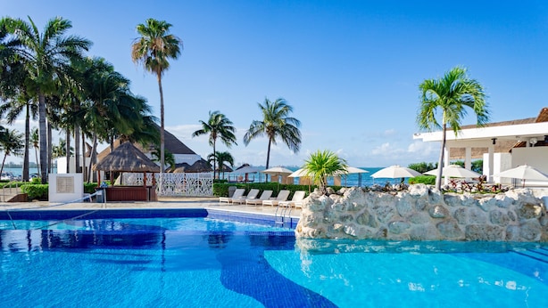 Ein klares, blaues Schwimmbecken mit Liegestühlen und Palmen, umgeben von einer Steinmauer und einem Gebäude mit einem Reetdach.