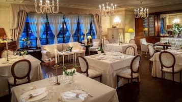 Una sala da pranzo formale con diversi tavoli apparecchiati per un pasto, eleganti lampadari e vista sull'esterno attraverso ampie finestre.