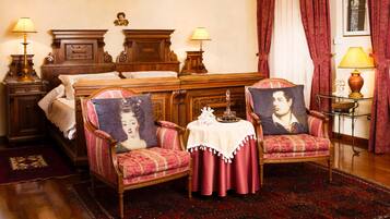 A traditional room with a bed, two armchairs with decorative cushions, a small table with a vase, and a red carpet.