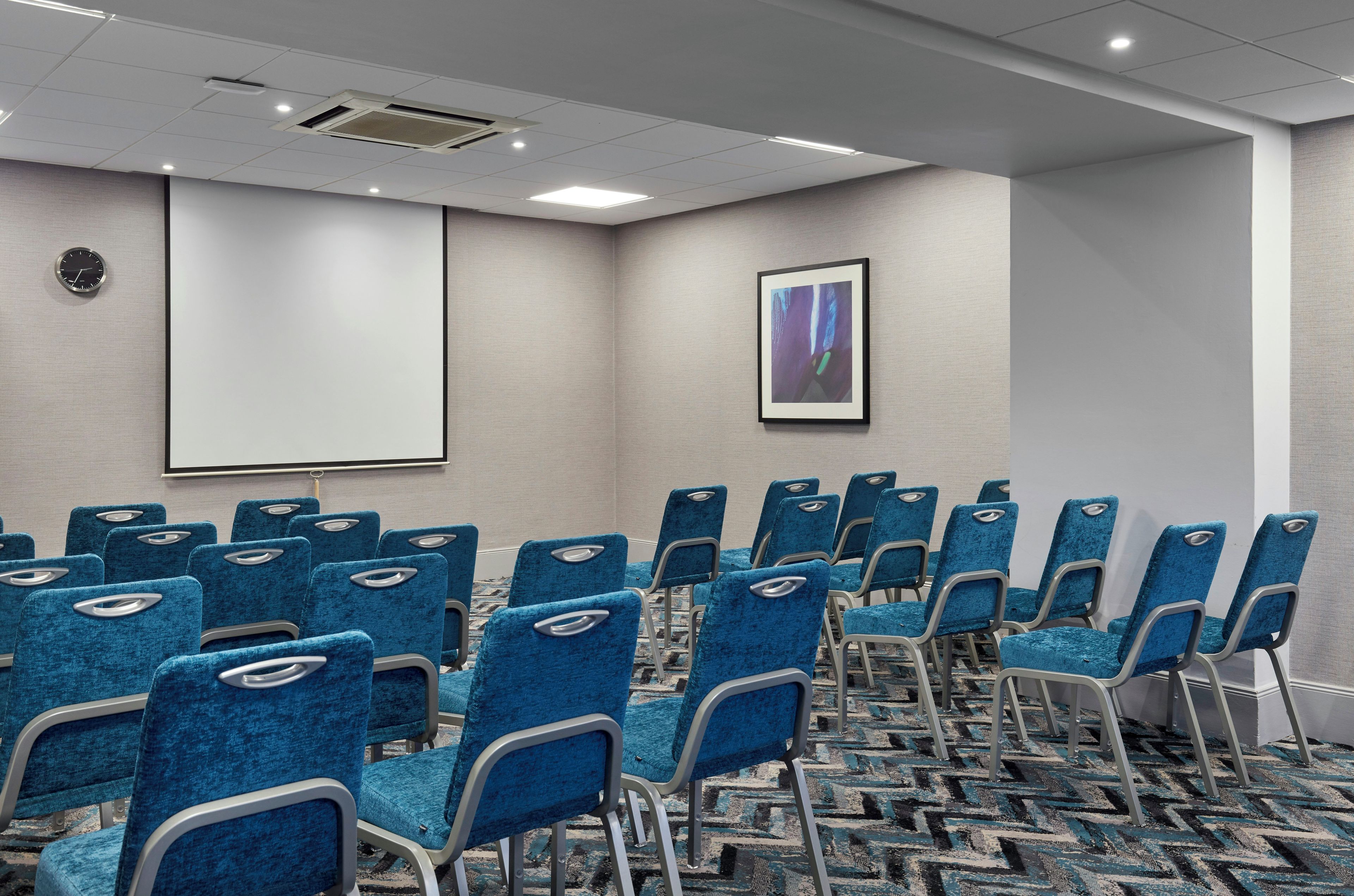 A conference room with rows of blue chairs, a projector screen, and a framed abstract painting on the wall.