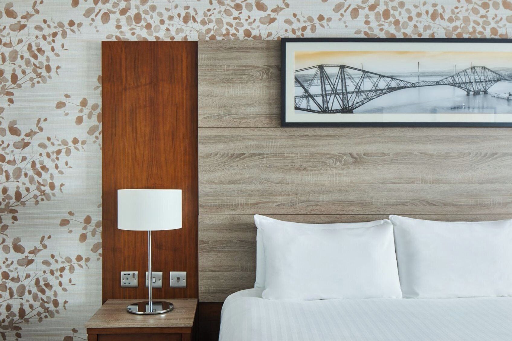 Habitación de hotel con cabecera de madera, cama blanca, mesita de noche con lámpara y un cuadro de un puente.