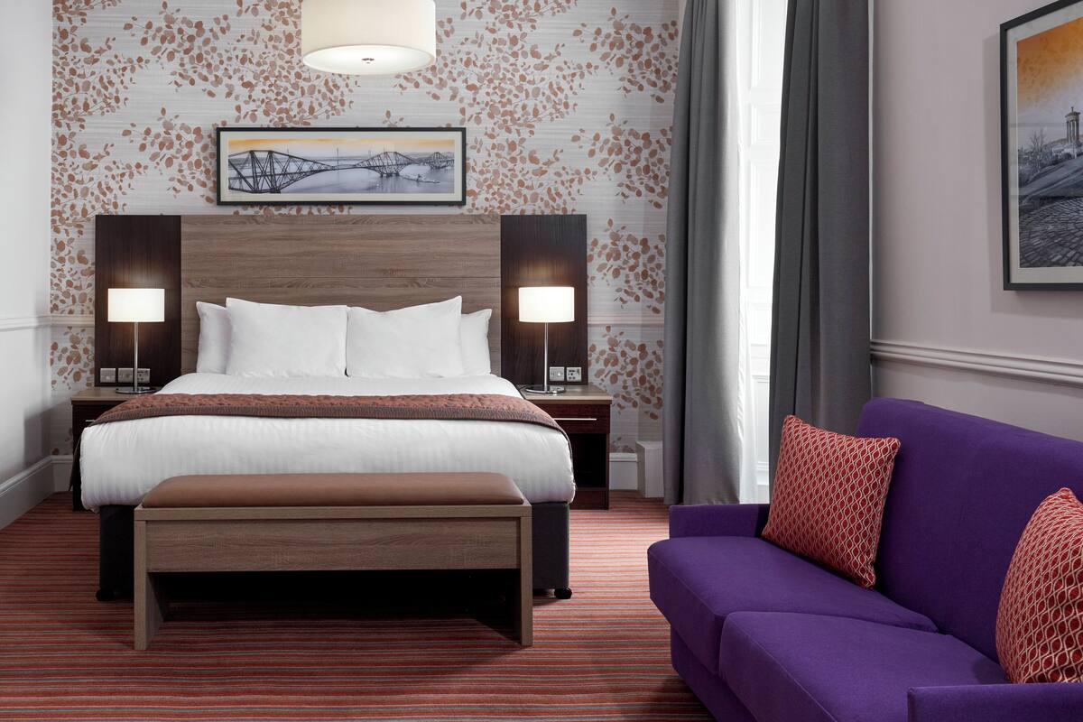 A hotel room with a large bed, two bedside lamps, a wooden headboard, a framed picture above the bed, a purple sofa with patterned cushions, and a red and white patterned pillow.