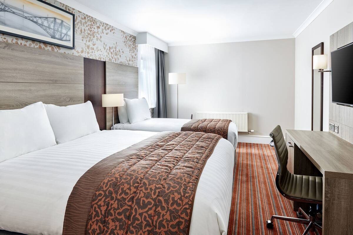 A hotel room with two beds, a desk, a chair, a TV, and a window with curtains.