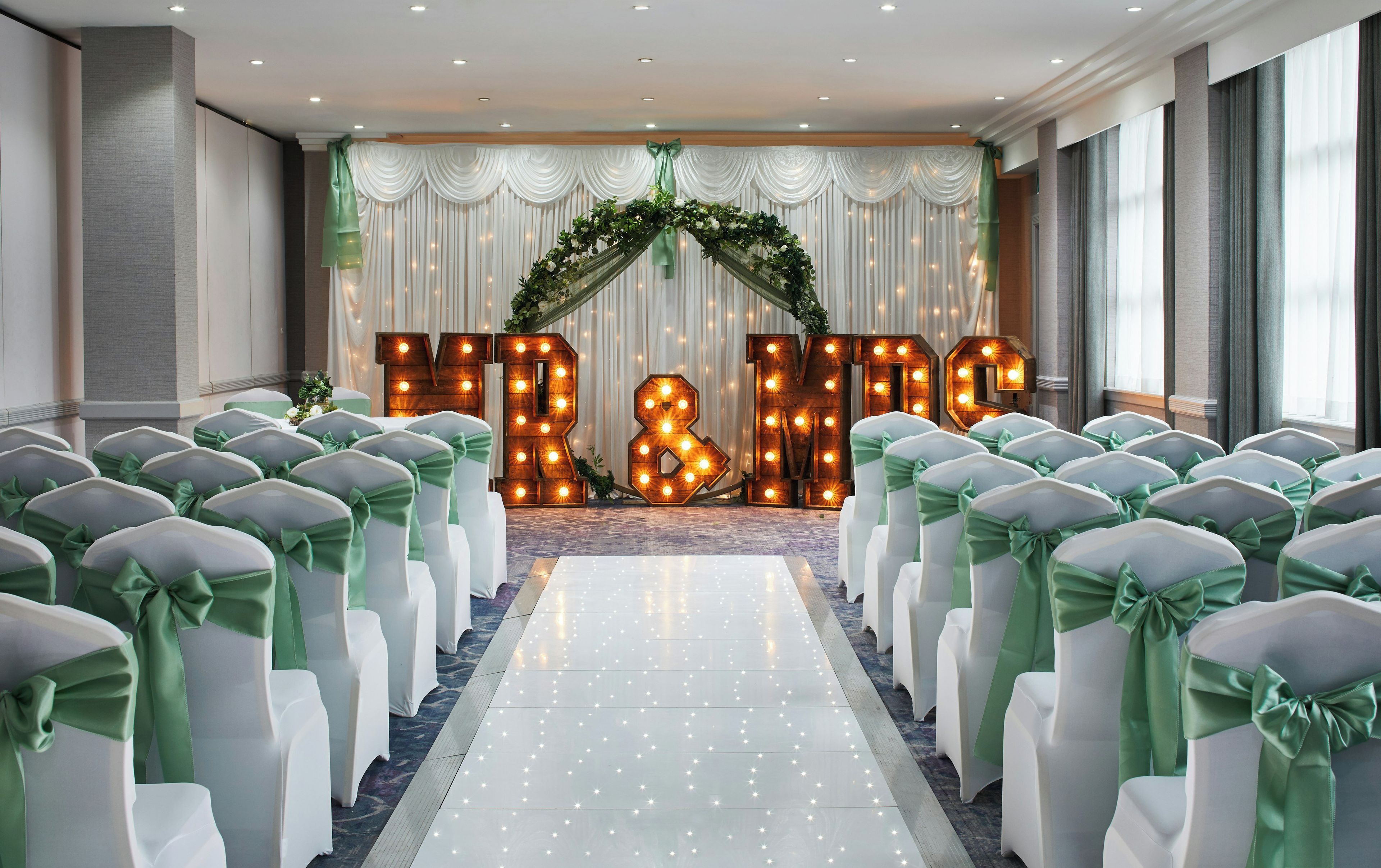 A wedding venue with rows of white chairs with green sashes, a floral arch, and illuminated 'MR & M' letters.
