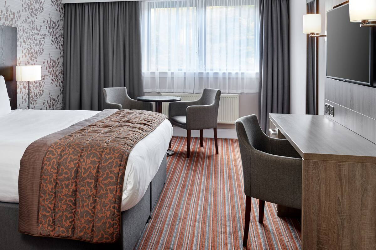 A hotel room with a large bed, a desk, two chairs, a television, and a window with curtains.