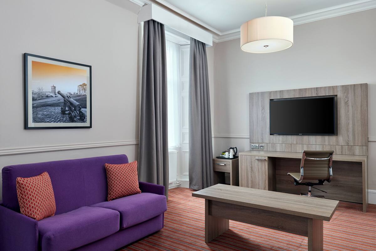 A modern hotel room with a purple sofa, a wooden coffee table, a flat-screen TV mounted on a wooden panel, and a window with grey curtains.