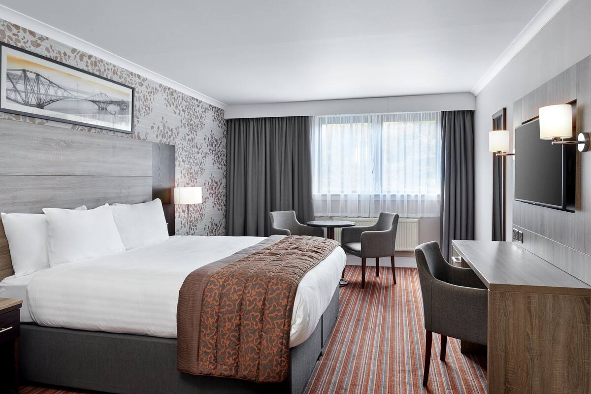 A hotel room with a bed, a desk, a chair, a TV, and a window with curtains.