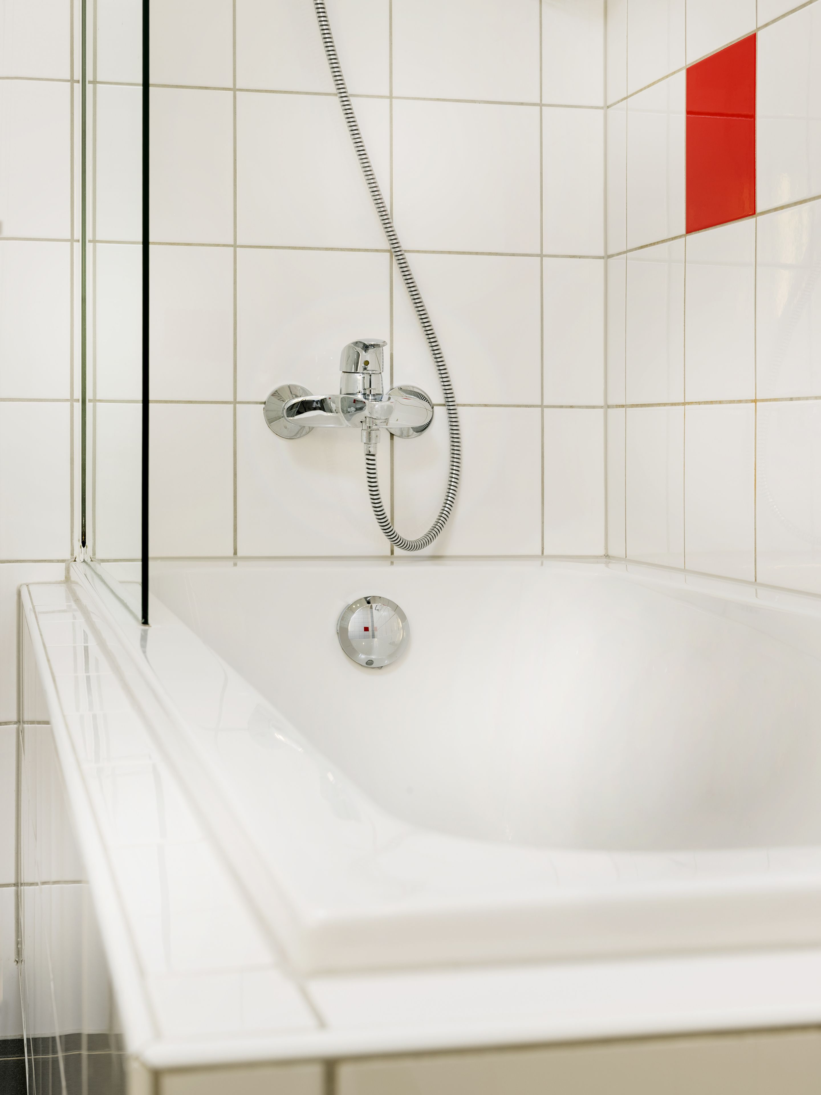 Een wit bad met een chromen kraan en douche, geplaatst tegen een betegelde muur met een rode accentkleur.