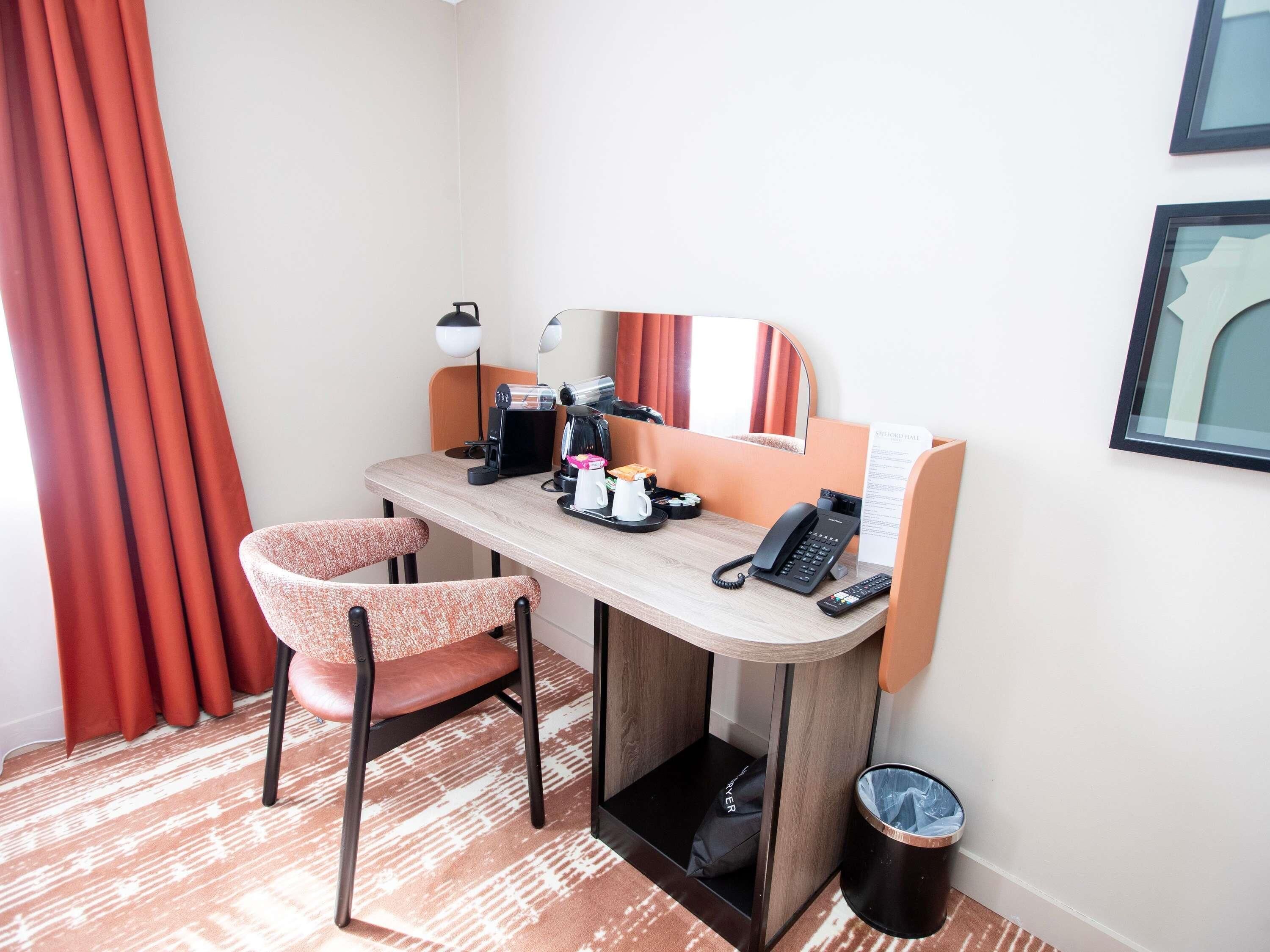 Quarto de hotel com escrivaninha, cadeira, cafeteira, telefone e lixeira.