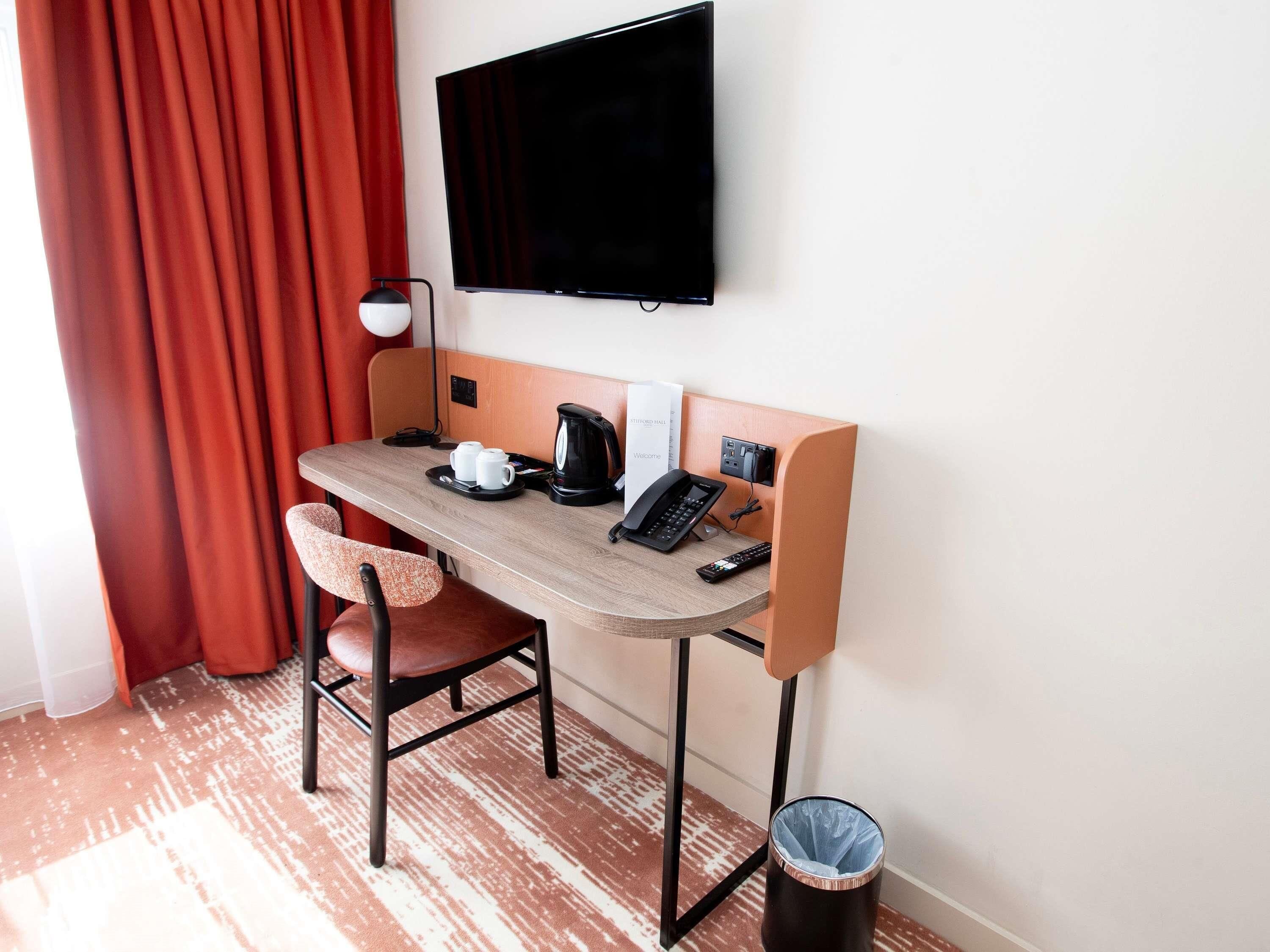 A hotel room with a desk, two chairs, a television, a coffee maker, and a trash can.