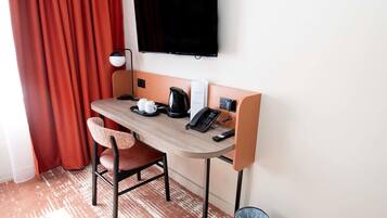 A hotel room with a desk, two chairs, a television, a coffee maker, and a trash can.