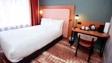 Habitación de hotel con cama, escritorio y silla. Hay una lámpara en el escritorio, un hervidor y un teléfono.