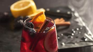 A glass of red beverage with ice, garnished with a slice of orange and a cinnamon stick, placed on a dark wooden surface.