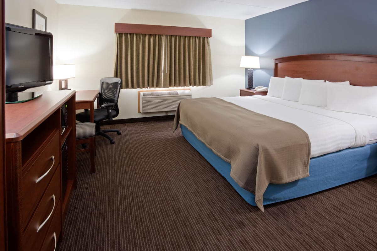 A hotel room with a bed, a desk with a television, a chair, an air conditioning unit, and a nightstand with a lamp.