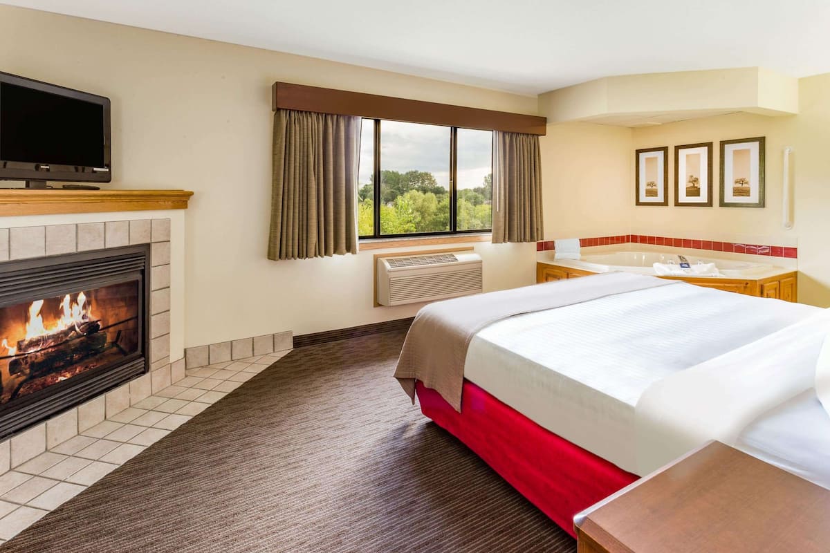 A hotel room with a fireplace, a bed, a TV, a window with curtains, and a bathroom with a bathtub.