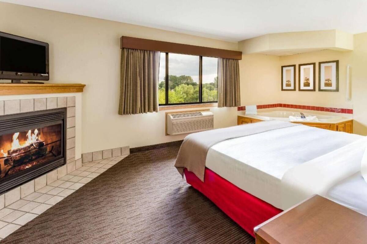 A hotel room with a fireplace, a bed, a television, a window with curtains, and a bathroom with a bathtub.