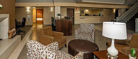 A spacious lobby with a high ceiling, a central seating area with patterned chairs, a brown sofa, and a wooden table with a lamp. A staircase leads to an upper level with a balcony.