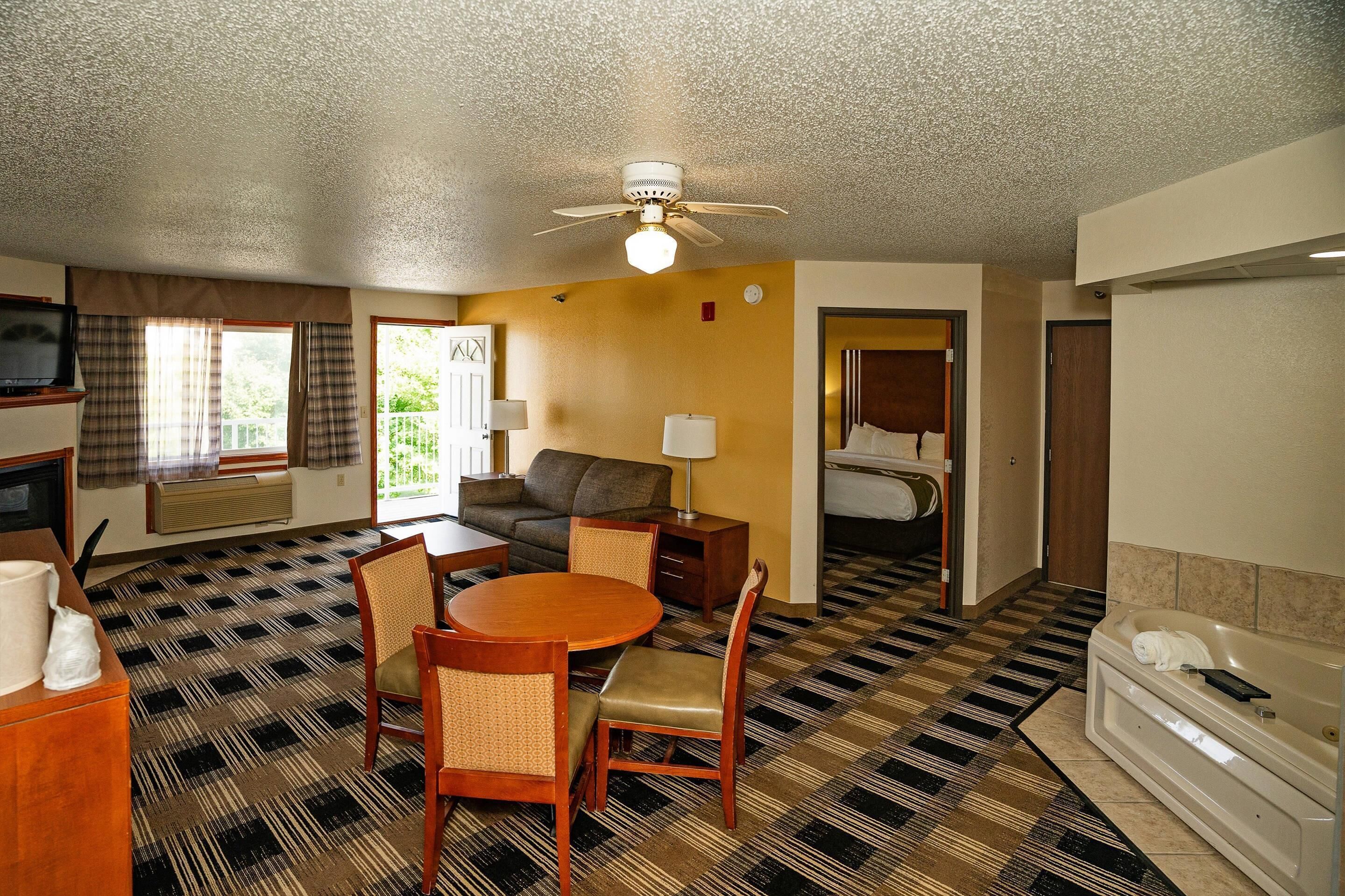 Quarto de hotel com chão xadrez, uma mesa de jantar com cadeiras, um sofá, uma TV, um ventilador de teto, um banheiro com banheira e uma janela com cortinas.