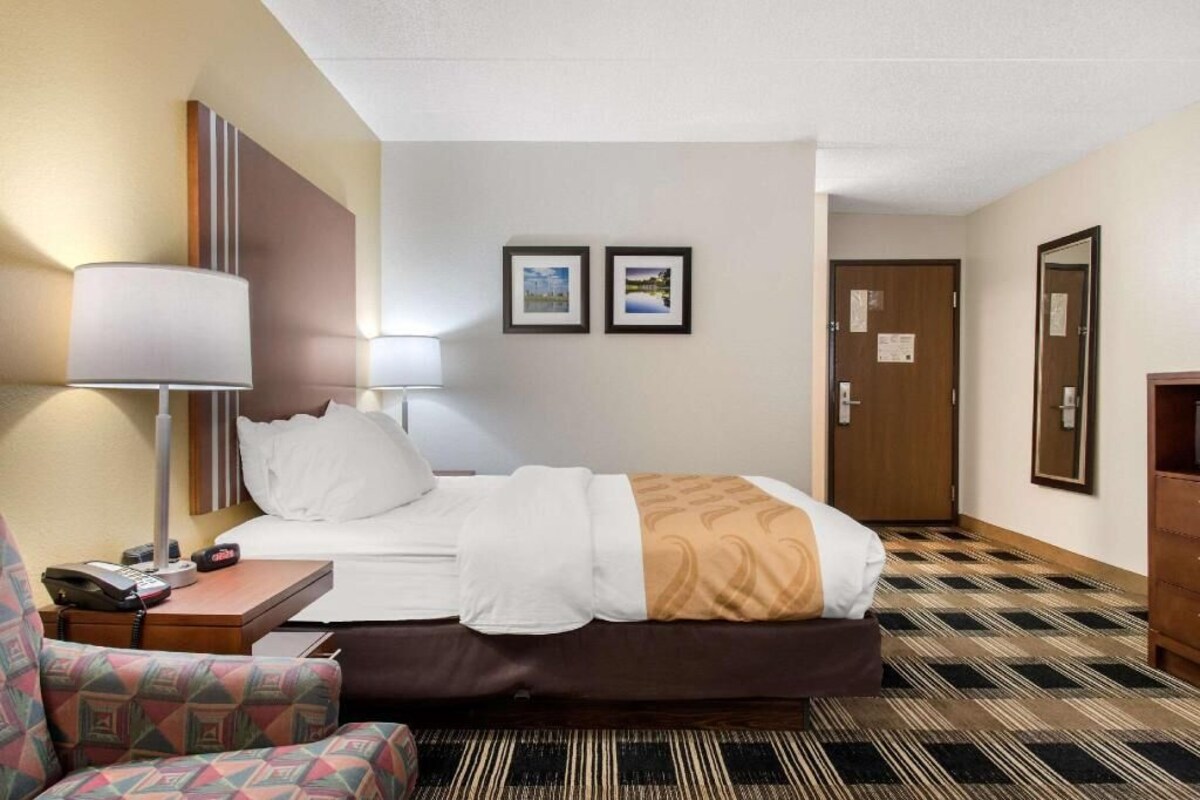 A hotel room with a bed, a chair, a desk with a lamp, a phone, and two framed pictures on the wall.