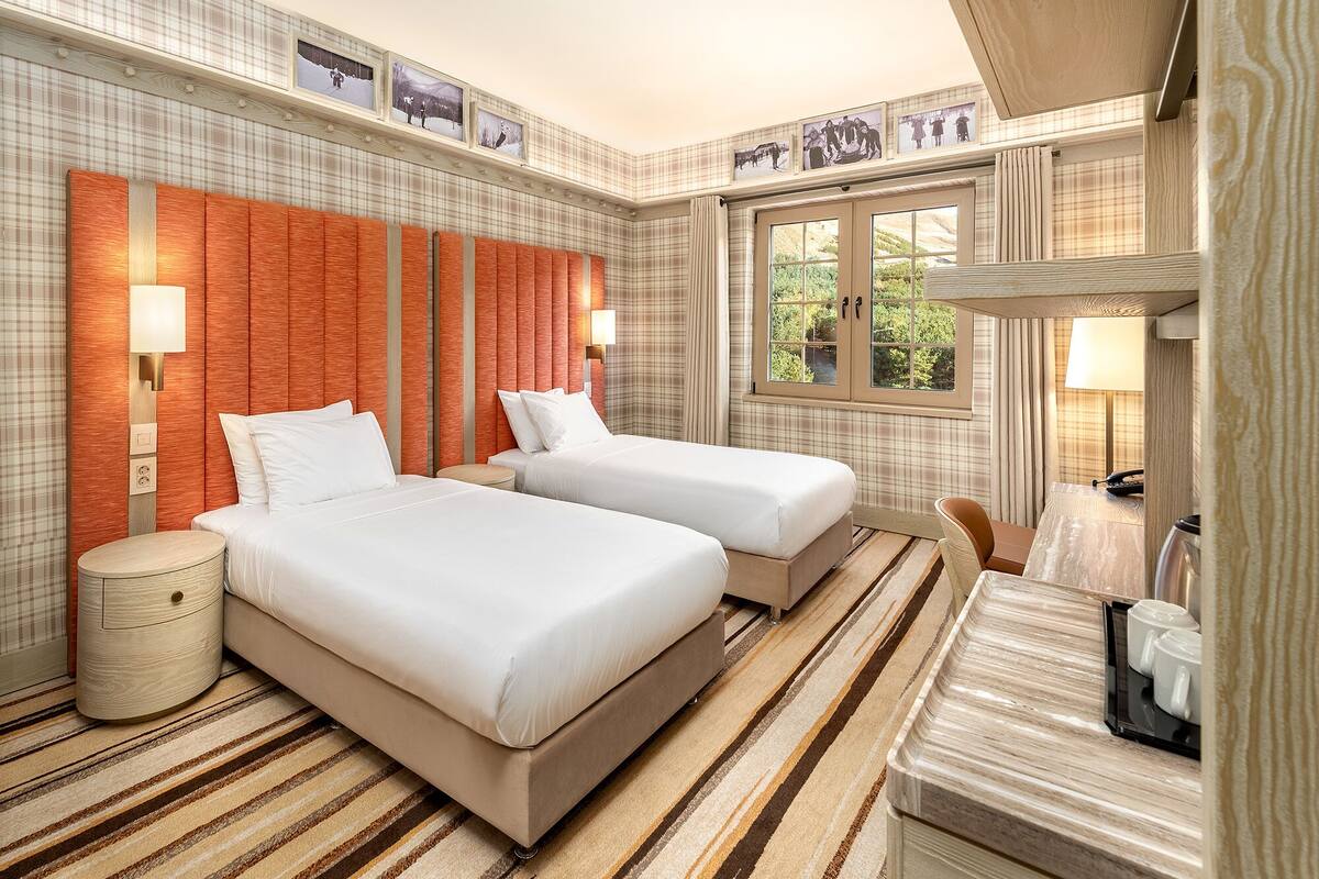 A hotel room with two beds, a large window, and a view of greenery outside.